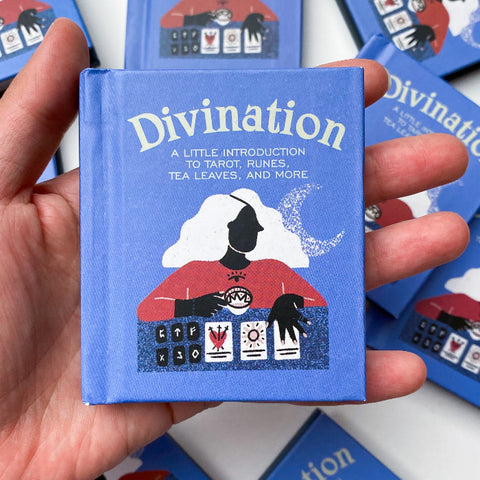 Hand holding a blue book titled 'Divination' with tarot card designs on a white background.