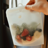 A hand holds a translucent silicone food storage bag with fruit inside, positioned in front of a refrigerator door.