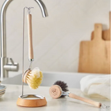 A wooden-handled dishwashing brush with a yellow bristle head hangs on a metal stand, while a second brush with dark bristles lies on the counter nearby. The scene is set in a bright kitchen with a sink, faucet, stacked plates, and a cutting board in the background.