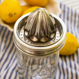 A stainless steel manual citrus juicer with a ribbed cone-shaped压榨 head, mounted on a clear glass mason jar. The jar sits on a blue and white striped cloth with fresh yellow lemons in the background.