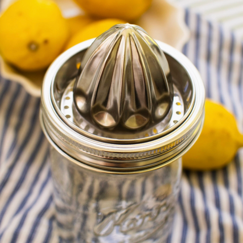 A stainless steel manual citrus juicer with a ribbed cone-shaped压榨 head, mounted on a clear glass mason jar. The jar sits on a blue and white striped cloth with fresh yellow lemons in the background.