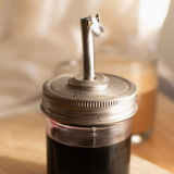 A small clear glass jar with a silver metal pour spout lid, filled with a very dark red liquid, set on a wooden surface with a softly blurred background.