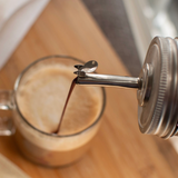 A close-up of a metal pour spout dispensing dark coffee into a glass mug on a wooden surface.