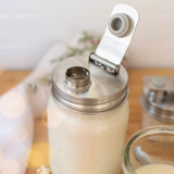 A glass jar with a stainless steel lid and a hinged flip-up straw cover, filled with a white beverage, set on a wooden surface with a softly blurred background.