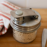 A small glass spice jar with a stainless steel lid and crank handle, filled with ground beige spice, set on a wooden cutting board next to a striped cloth and a knife.