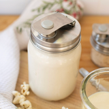 A stainless steel milk pitcher with a flip-top lid sits on a wooden table next to a glass of milk, with popcorn and a floral cloth in the background.