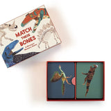 A boxed card game titled "Match These Bones" shown on a white background. The open box contains two illustrated cards featuring dinosaur skeletons and a bird-like creature, with the matching box lid placed above showing additional dinosaur skeleton illustrations and the title text.