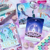 A styled flat-lay of the 'Work Your Light Oracle Cards' deck by Rebecca Campbell, showing the colorful card box, several illustrated oracle cards, crystals, and a crochet blanket on a pink watercolor background.