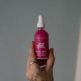 Hand holding a pink bottle labeled 'Rose Dew' against a gray background