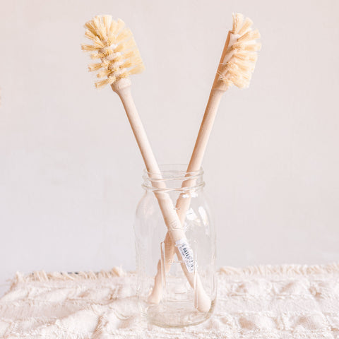 Two wooden brushes with natural bristles, designed for cleaning drinking bottles, standing upright in a glass bottle.