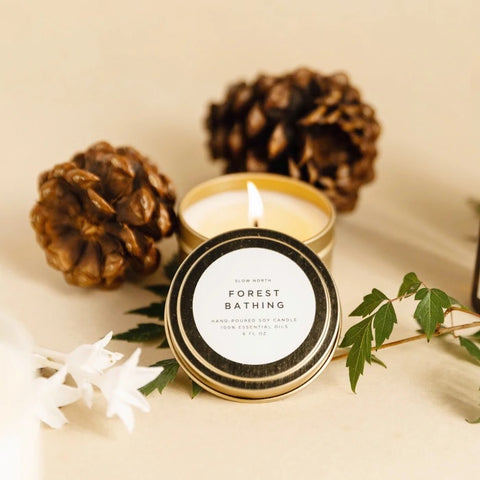 A small soy wax candle with a lit flame, surrounded by pine cones and greenery, with a label that reads 'FOREST BATHING'.