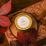 A small gold jar candle labeled 'Cabin Retreat' by Slow North, surrounded by autumn-themed decorations like leaves and a wooden texture.
