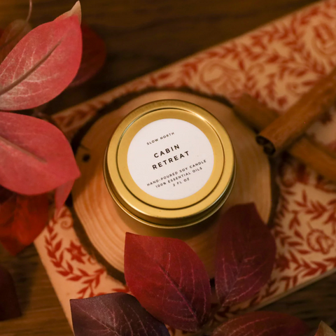 A small gold jar candle labeled 'Cabin Retreat' by Slow North, surrounded by autumn-themed decorations like leaves and a wooden texture.